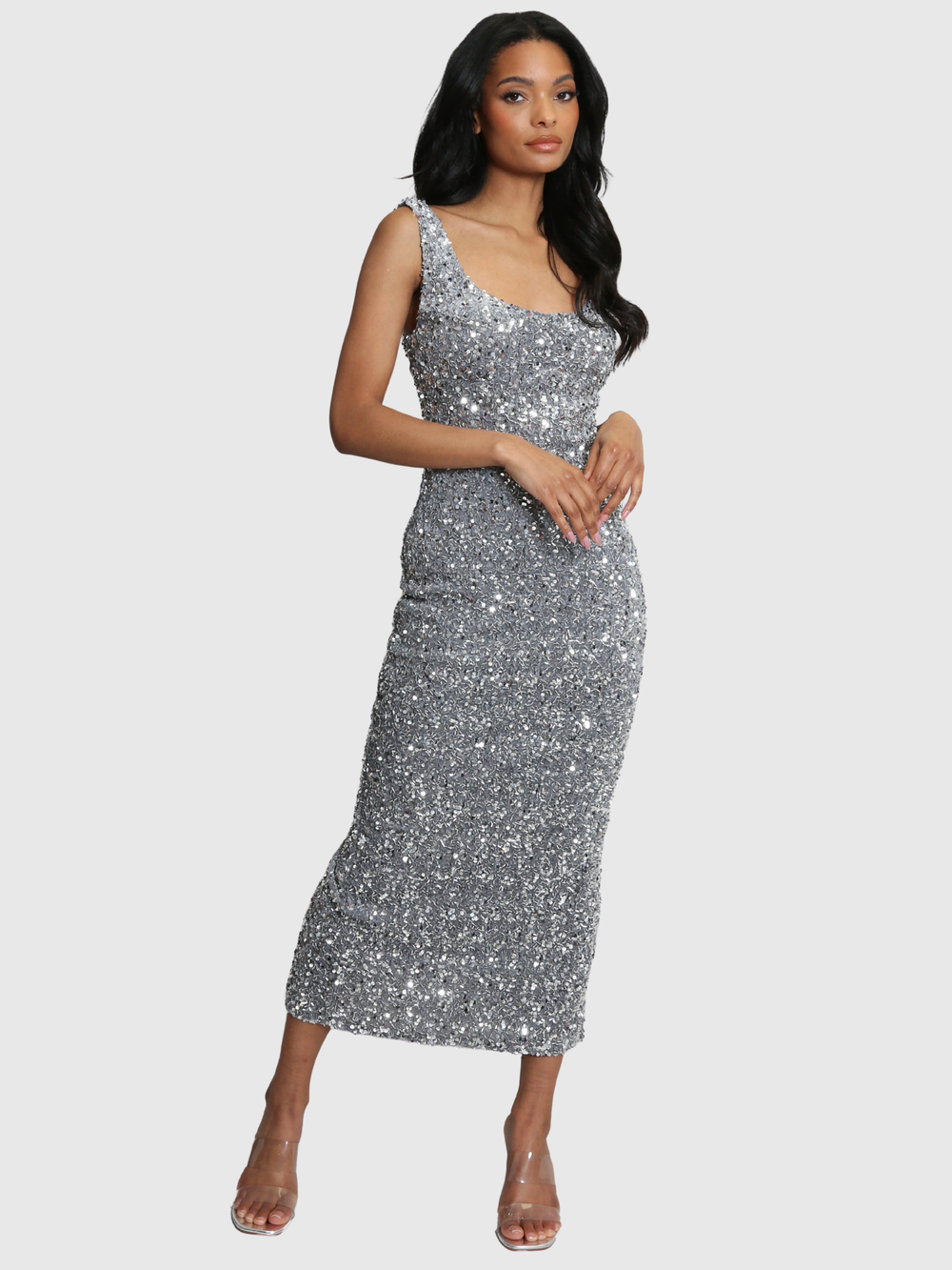Full shot. Model weras a deluxe silver sequin midi dress with a square neckline and zip back fastening. Model has brown skin and long black hair. Model faces the camera and places her hands in front, she stares into the camera. 