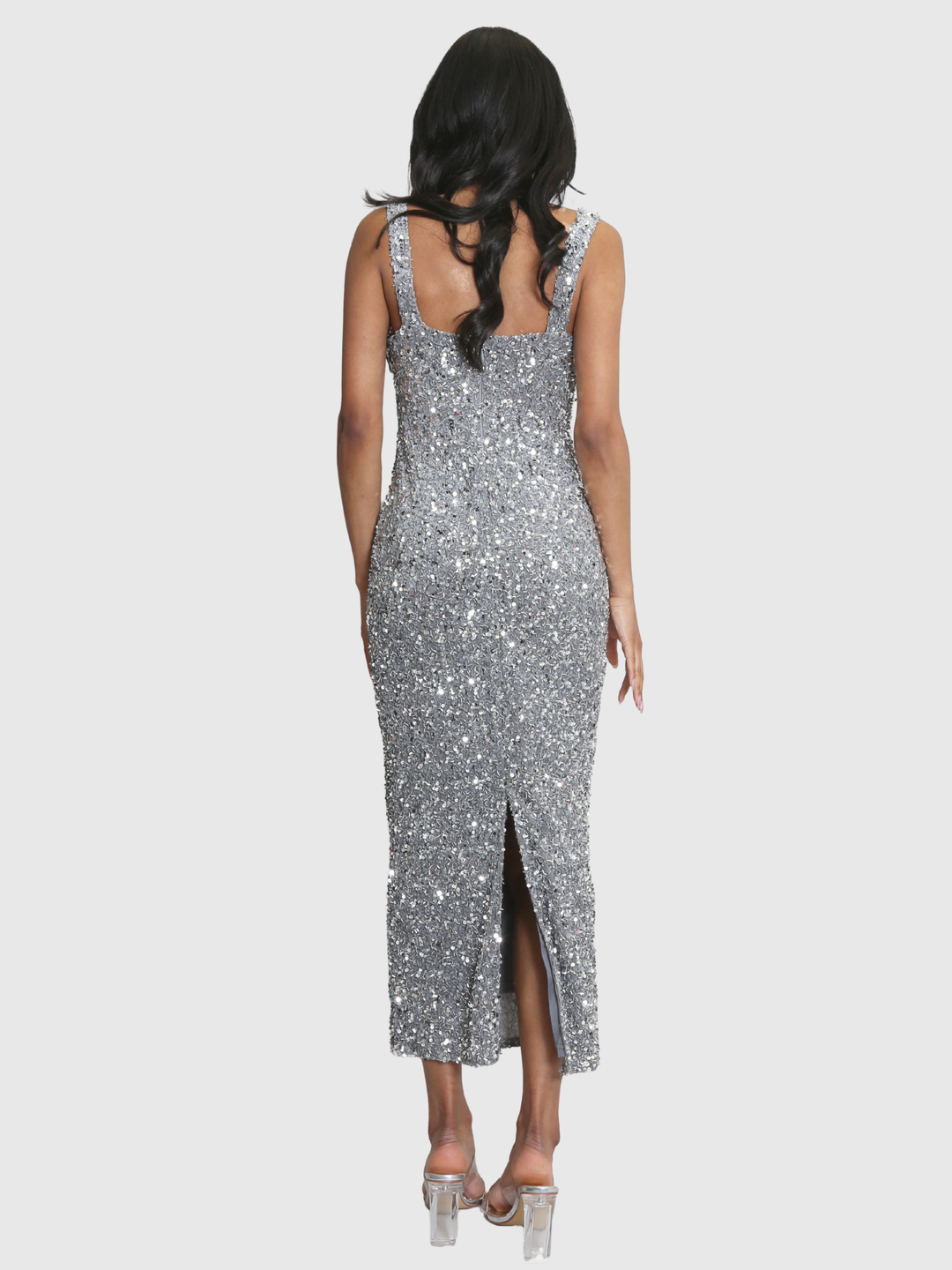 Full shot. Model weras a deluxe silver sequin midi dress with a square neckline and zip back fastening. Model has brown skin and long black hair. The model stands with her back to the camera, the back of the dress can be seen, including the bottom slit. 