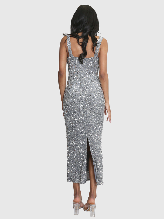 Full shot. Model weras a deluxe silver sequin midi dress with a square neckline and zip back fastening. Model has brown skin and long black hair. The model stands with her back to the camera, the back of the dress can be seen, including the bottom slit. 