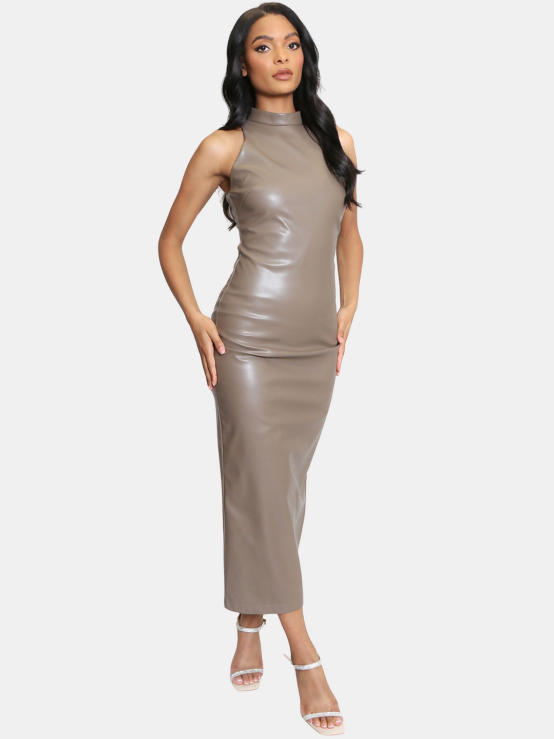 Full length shot. Female model stands in front of a light taupe plain background. The model wears a taupe faux leather midi dress, with a high neck and sleeveless design. The model has light model stands with her hips brown skin and long black hair. The model places her hands on her hips. 
