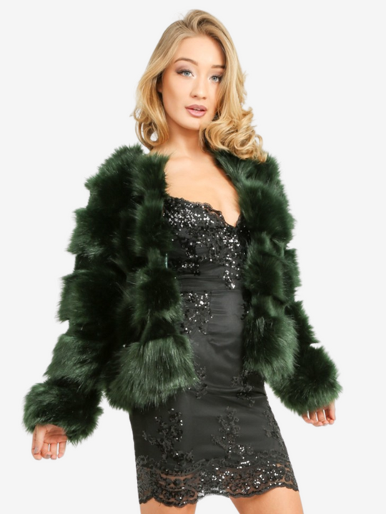 Model wears a soft faux fur long sleeve green jacket. The jacket has a front fastening and  satin like inner lining. Model wears the jacket open, she is wearing a  black sequin dress.  Model stands slighly to the side with her hands by her sides. Model faces the camera. 