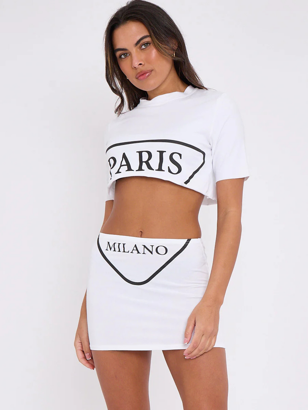 Model wears a Paris Milano Crop Top and matching skirt, which has the graphic t shirt at the front of both the crop top and skirt. The model has tanned skin and  long brown hair, she stands slightly tilted to the side and faces the camera, she places her hands by  her sides. 