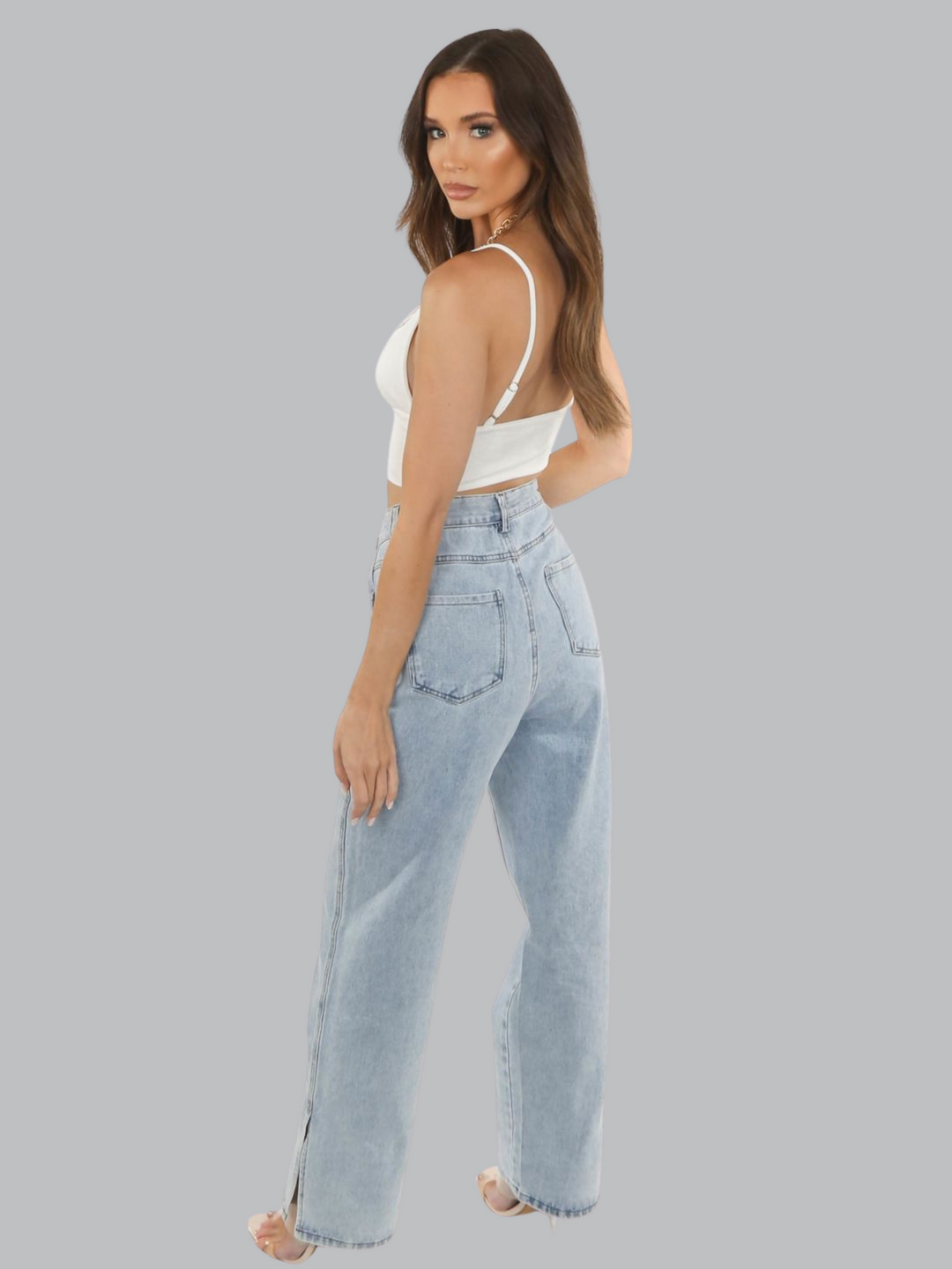 Full length shot. Model wears a white slinky v neck bralette, with adjustable straps and  a thick  white band at the waist. Model has brown hair, she stands with her back slightly to the the camera, showing her bralette and the denim jeans, she turns her head to look into the camera.
