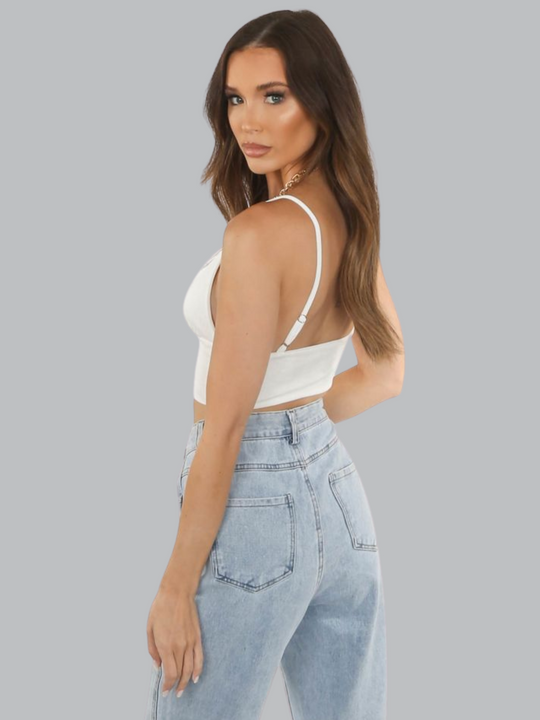 Model wears a white slinky v neck bralette, with adjustable straps and  a thick  white band at the waist.  Model stands to the side, so that the back of her jeans and the bralette can be seen. The model turns her head so that she looks directly into the camera.