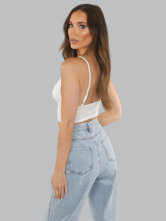 Mid length shot. Model wears a white slinky v neck bralette, with adjustable straps and  a thick  white band at the waist. Model stands to the side so that her back can be see, she turns her head to look at the camera