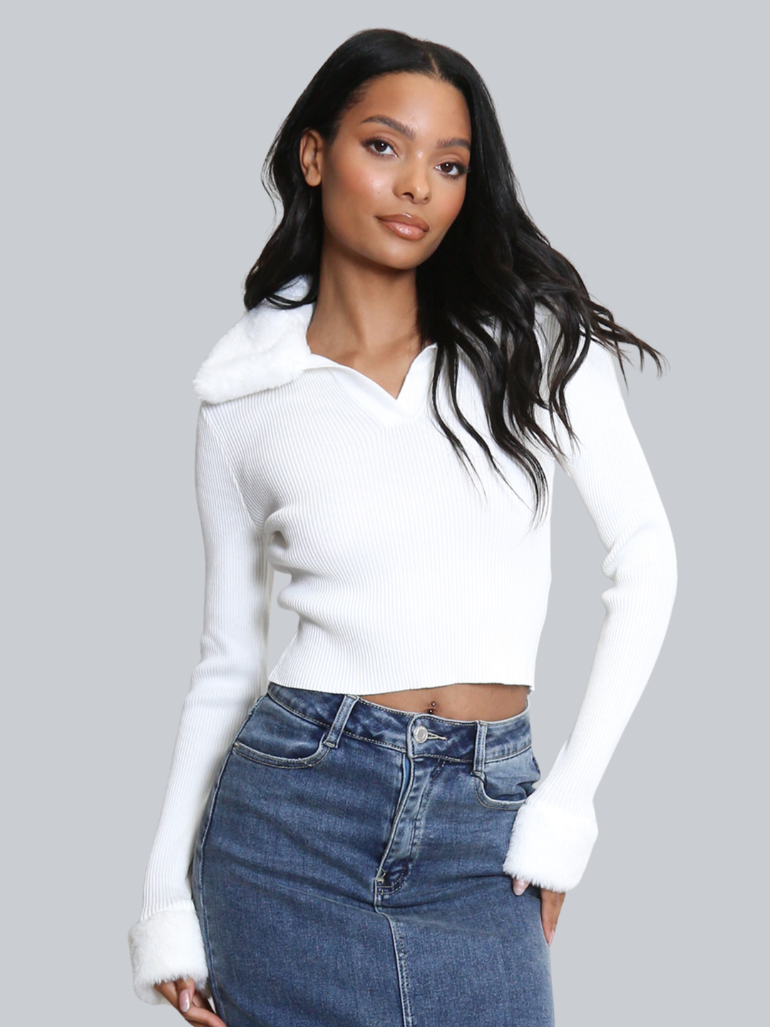 Female model stands in front of a plain background. The model wears a white ribbed jumper with faux fur sleeved cuffs and faux fur collar. The faux fur jumper has a  v neck front. The female model has light brown skin and long black hair, she looks into the camera and tilts her head slightly, she places one hand on her hip and the other by her side. 