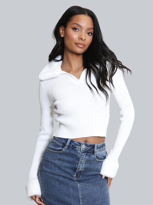 Female model stands in front of a plain background. The model wears a white ribbed jumper with faux fur sleeved cuffs and faux fur collar. The faux fur jumper has a  v neck front. The female model has light brown skin and long black hair, she looks into the camera and tilts her head slightly, she places one hand on her hip and the other by her side. 