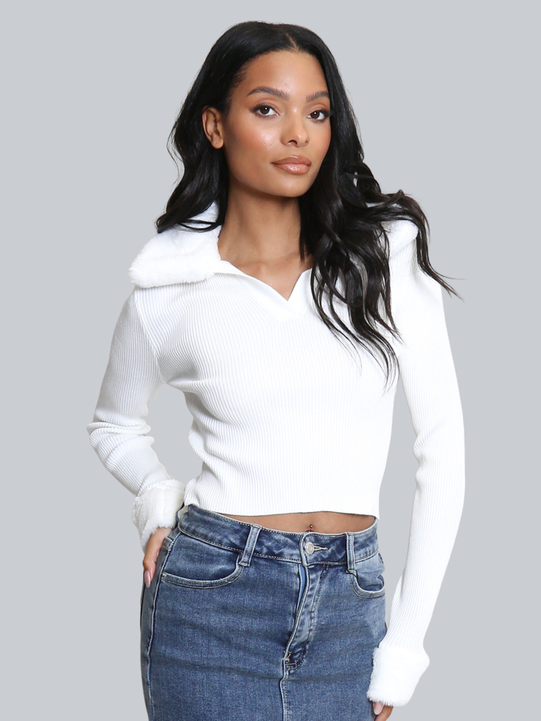 Female model stands in front of a plain background. The model wears a white ribbed jumper with faux fur sleeved cuffs and faux fur collar. The faux fur jumper has a  v neck front. The female model has light brown skin and long black hair, the model faces forward , placing one hand on her hip and the other by her side. 