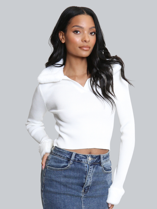 Female model stands in front of a plain background. The model wears a white ribbed jumper with faux fur sleeved cuffs and faux fur collar. The faux fur jumper has a  v neck front. The female model has light brown skin and long black hair, the model faces forward , placing one hand on her hip and the other by her side. 