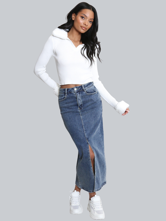 Full length shot. Female model stands in front of a plain background. The model wears a white ribbed jumper with faux fur sleeved cuffs and faux fur collar. The faux fur jumper has a  v neck front. The female model has light brown skin and long black hair, the model faces the camera and wears a long denim skirt and chunky trainers. 