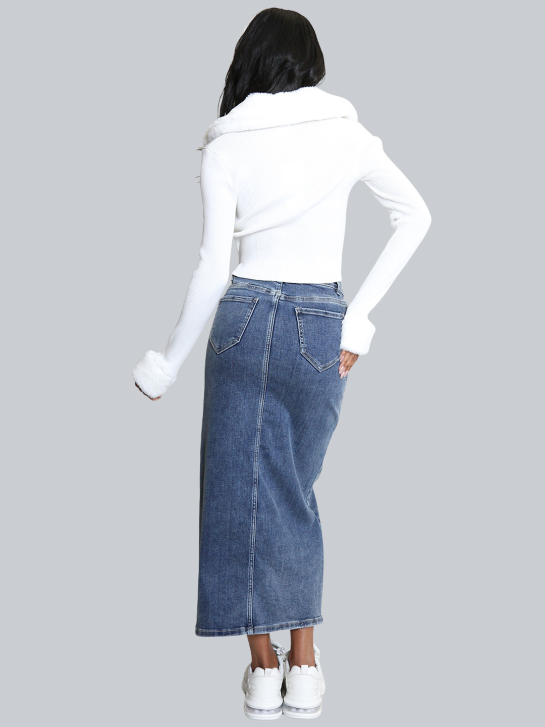 Full length shot. Female model stands in front of a plain background. The model wears a white ribbed jumper with faux fur sleeved cuffs and faux fur collar. The faux fur jumper has a  v neck front. The female model has light brown skin and long black hair, the model has her back to the camera, she places one hand on her hip and the other by her sides, the faux fur collar can be seen. 