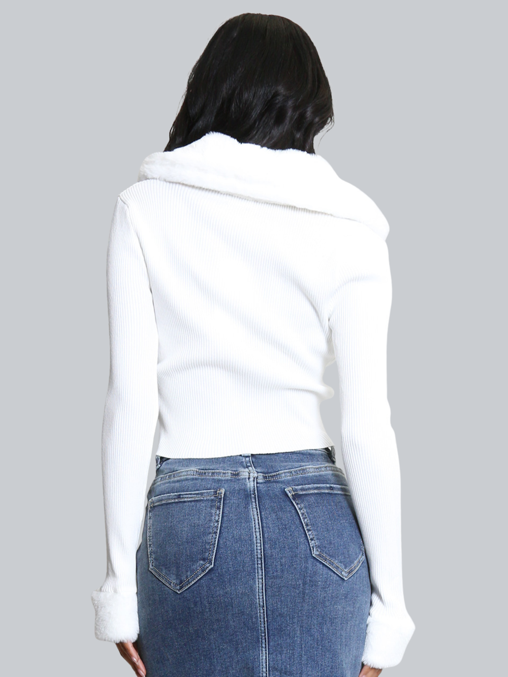 Female model stands in front of a plain background. The model wears a white ribbed jumper with faux fur sleeved cuffs and faux fur collar. The faux fur jumper has a  v neck front. The female model has light brown skin and long black hair, she stands with her back to the camera, placing her hands by her sides, the faux collar and cuffs can be seen. 