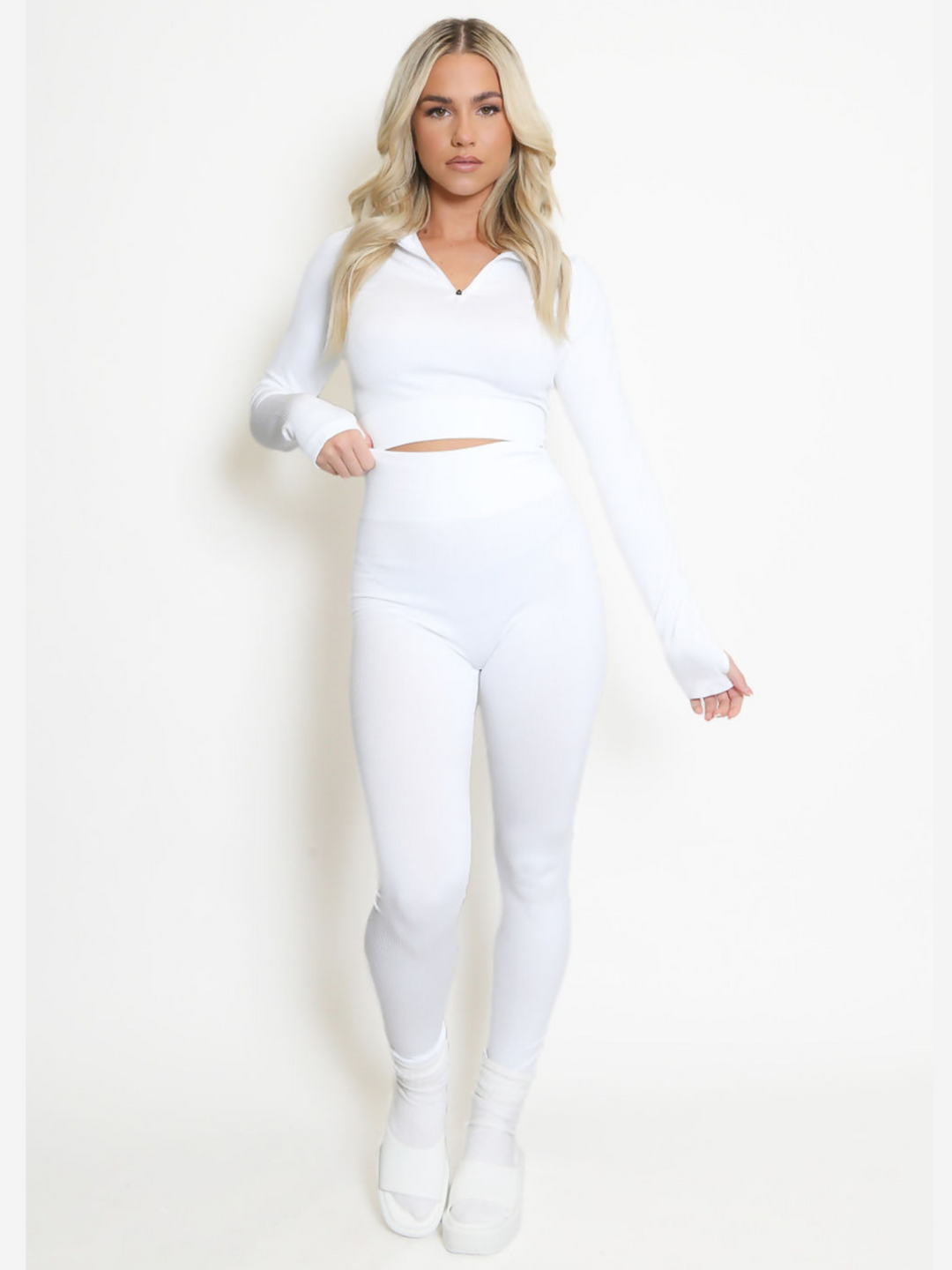 Full length shot. Female model wears a white co-ordinated set, she stands before a plain cream background. The model wears a long sleeve ribbed crop top and front zip fastening. The model wears a matching ribbed leggings. The model has tanned skin and long blonde hair, she stands with one leg slightly bent, faces the camera and laces one hand by her side and the other on her hip. 