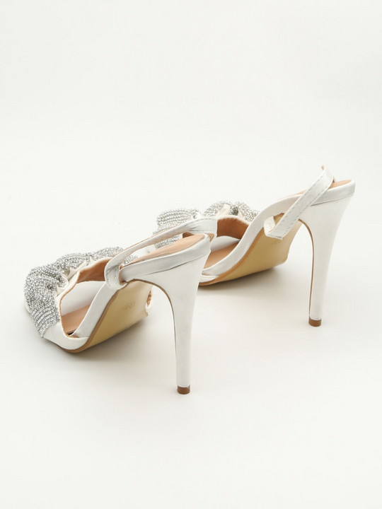 Pair of white high-heeled shoes with embellishments on a light background. The shoe is placed so that the high heel is closet to the camera and the diamante embelished strap furthest away. 
