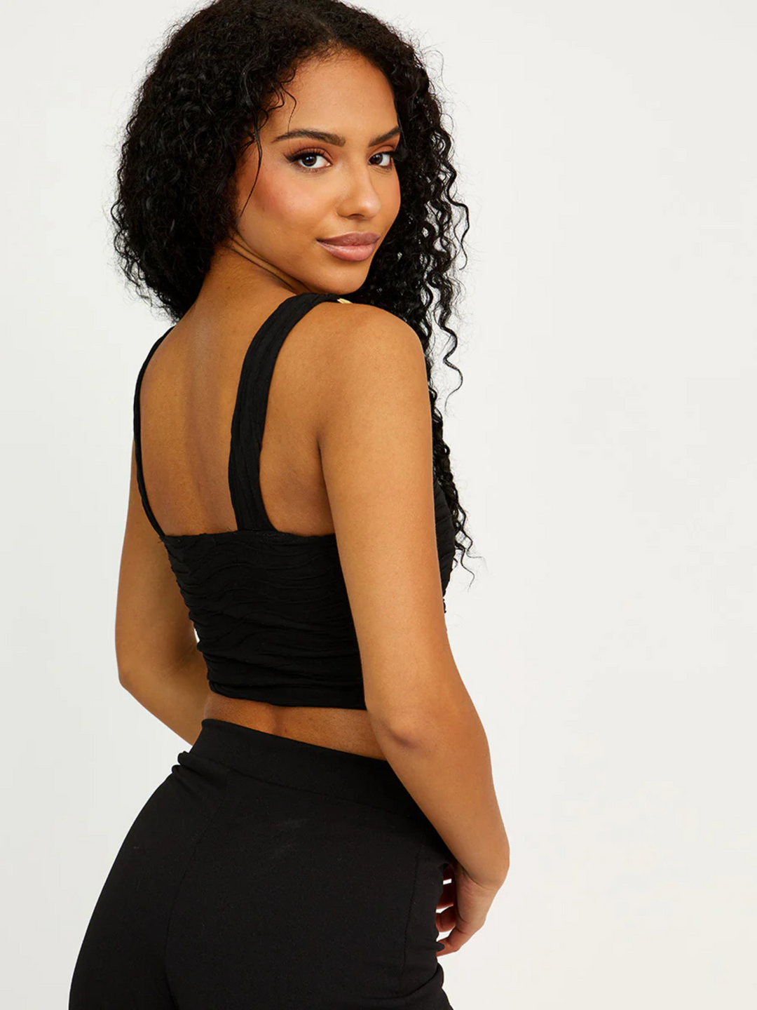 Model wears a black textured crop top that has a square neckline and chunky gold detailing at the straps. Model has light brown skin with curly long black hair, she stands with her side and the back of the top can be seen, she turns her head to look directly into the camera. 
