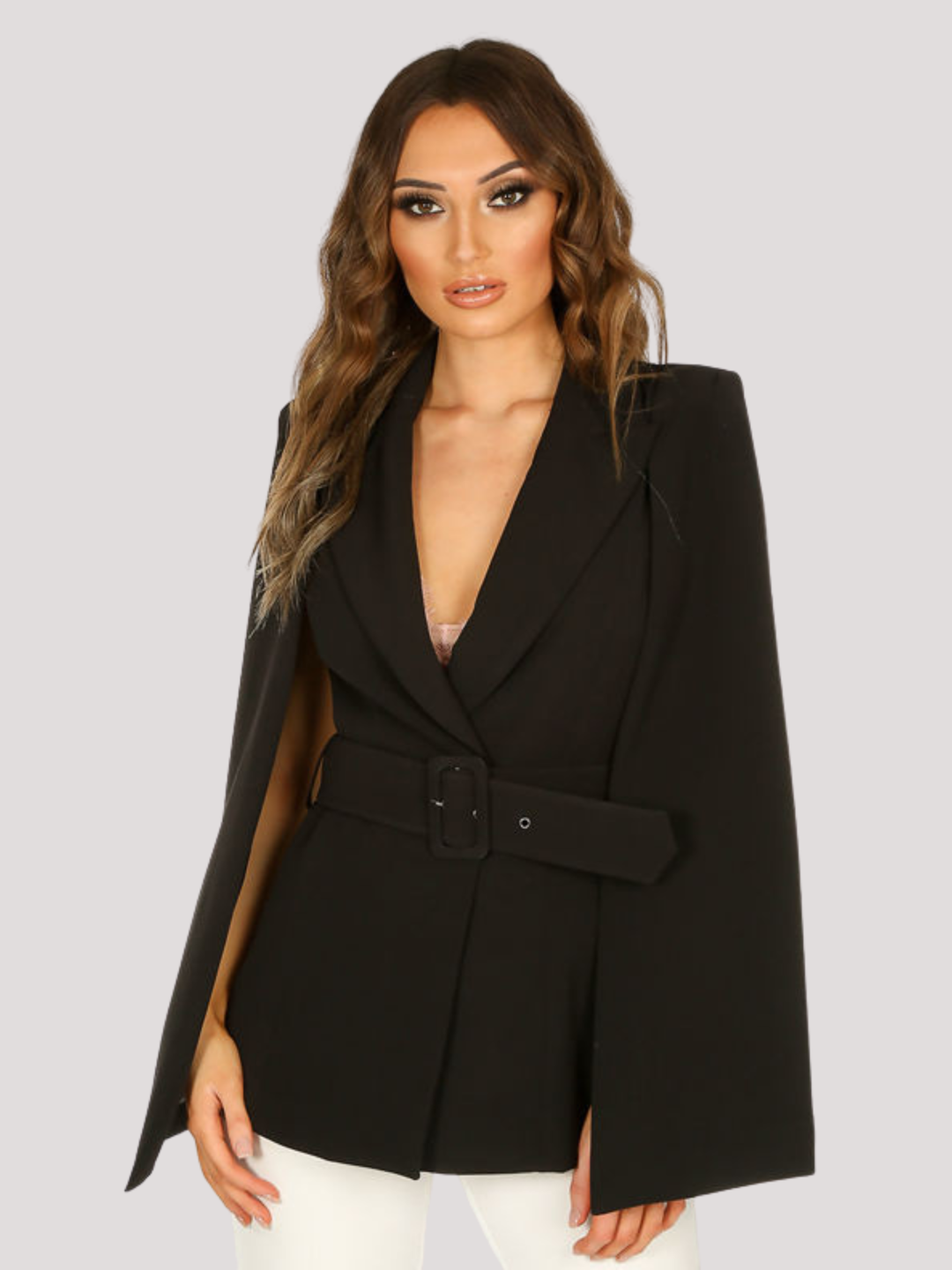 Model stands in front of a plain background. The model wears a tailored luxe blazer, with a bat cape shoulders. The black blazer has a v neck design and a belted waist. The model has tanned skin and long light brown hair, she faces forward and places her hands by her sides.