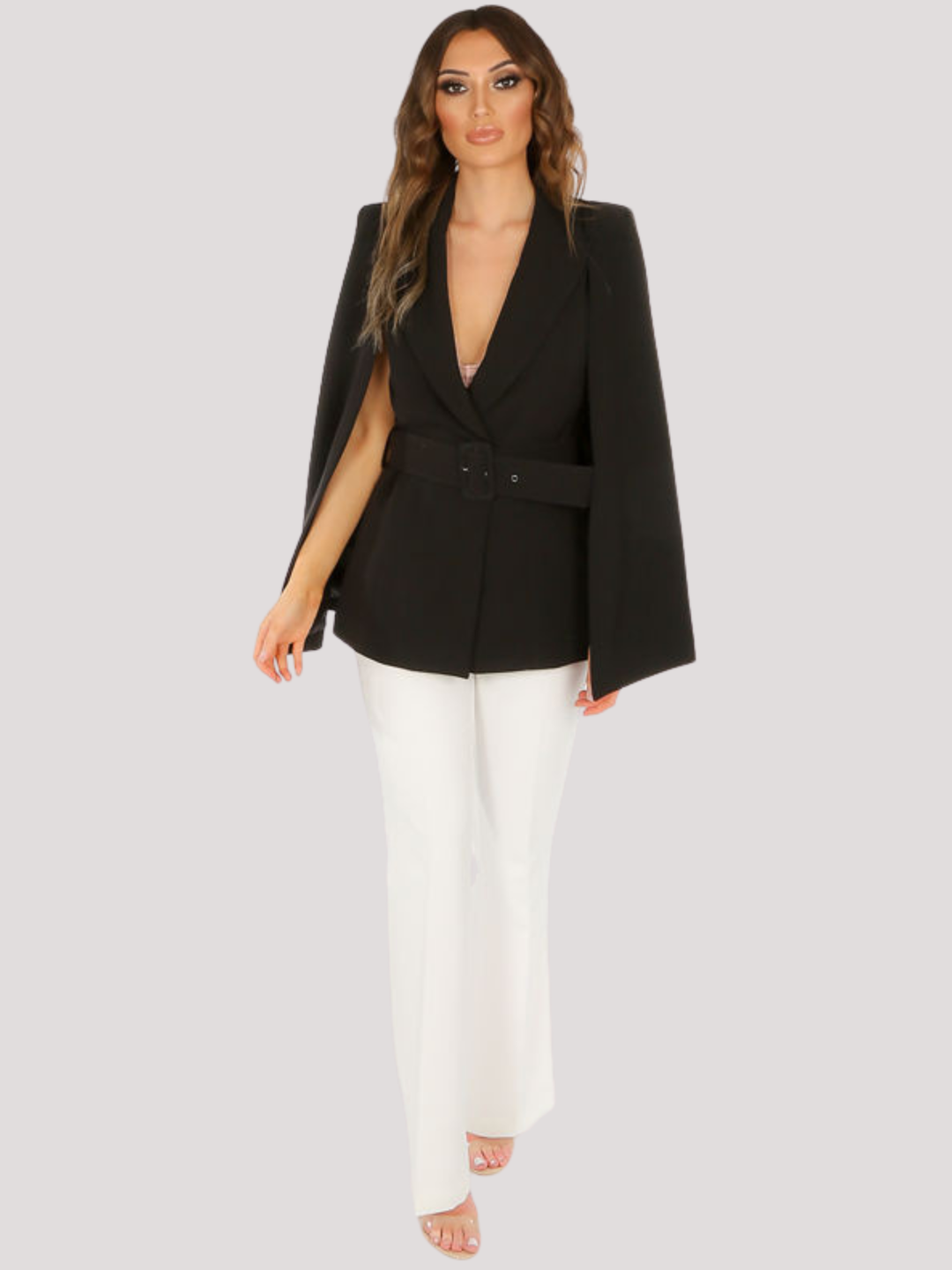 Full length shot. Model stands in front of a plain background. The model wears a tailored luxe blazer, with a bat cape shoulders. The black blazer has a v neck design and a belted waist. The model has tanned skin and long light brown hair, she faces the camera and has her hands by her sides. The model wears a white trousers and clear perspex heels.