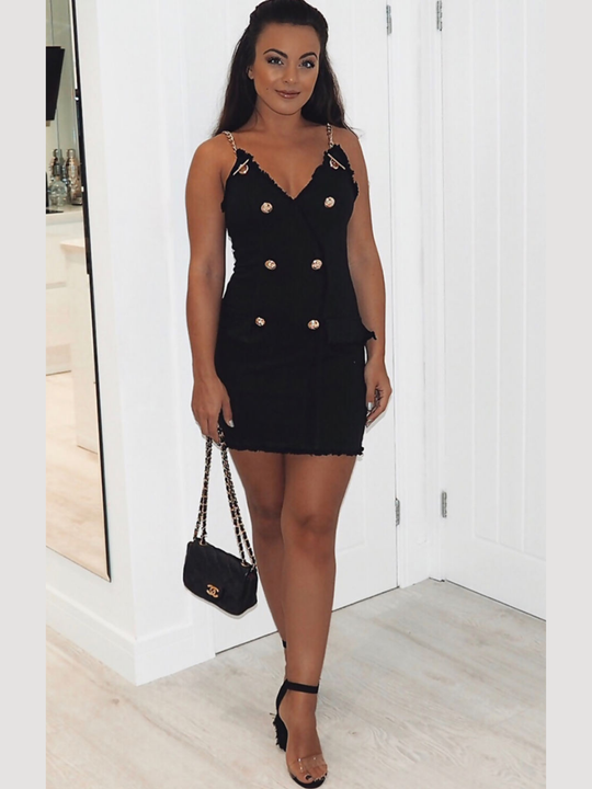 Female wears  a black denim mini dress, with wrap front bodycon design, front gold button detailing and gold chain straps and exposed zip design, she faces the camera and holds a black bag. 