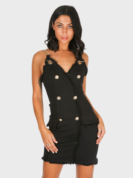 Model stands in front of a plain background. The model wears  a black denim mini dress, with wrap front bodycon design, front gold button detailing and gold chain straps and exposed zip design. The model has tanned skin and black hair, she faces the camera placing one hand on her hip and the other on her thigh. 