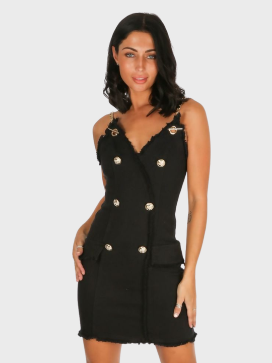 A female stands in front of  a plain background, she wears a black denim mini dress, with wrap front bodycon design, front gold button detailing and gold chain straps and exposed zip design. The model has tanned skin and black hair, she faces the camera placing one hand on her hip and the other on her thigh.