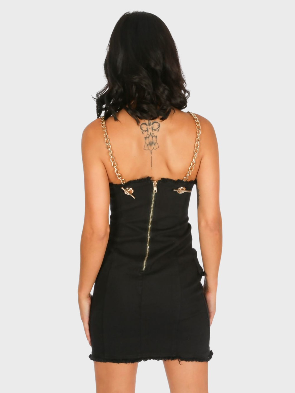 Model stands in front of a plain background. The model wears  a black denim mini dress, with wrap front bodycon design, front gold button detailing and gold chain straps and exposed zip design. The model has tanned skin and black hair, she stands with her back to the camera, the back of the dress can be seen. The exposed zip and chain straps can be seen. 