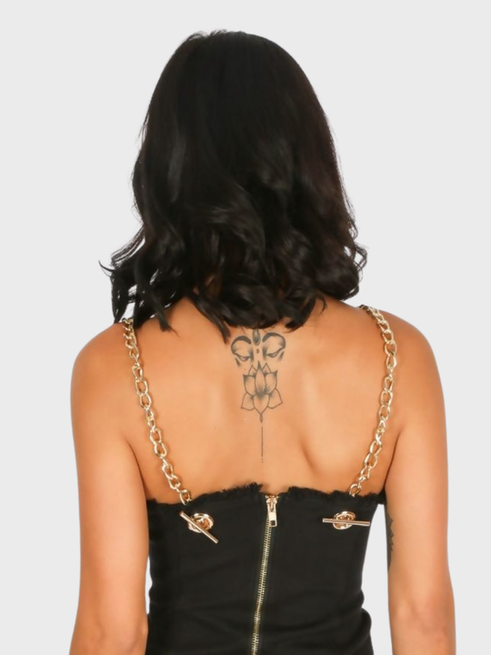 The model has tanned skin and black hair, she stands with her back to the camera, she wears a black mini dress, the  back of the black mini dress can be seen, including the exposed zip back and gold chain shoulder straps.  