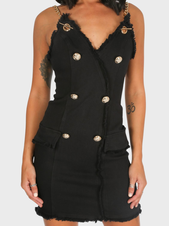 Close up. Model stands in front of a plain background. The model wears  a black denim mini dress, with wrap front bodycon design, front gold button detailing and gold chain straps and exposed zip design. 
