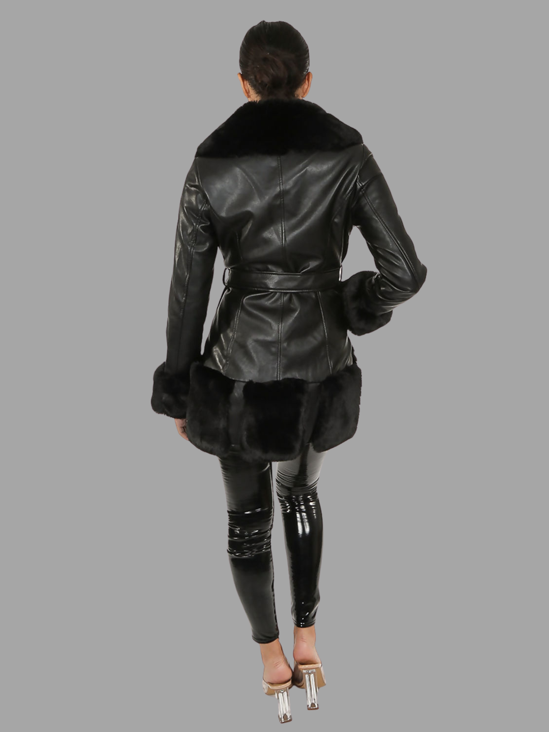 Full length shot. Female model stands in front of a plain grey background. The model wears a black faux leather coat with black faux fur lapel, black faux fur trim and black faux cuffs. The belt has a a faux leather belted waist. The model has tanned skin and black hair that is slicked back. The model stands with her back to the camera, the back of the jacket can be seen including faux fur trim, faux fur collar and sleeved faux fur cuffs. The model wears pu leather leggings and muled heels. 