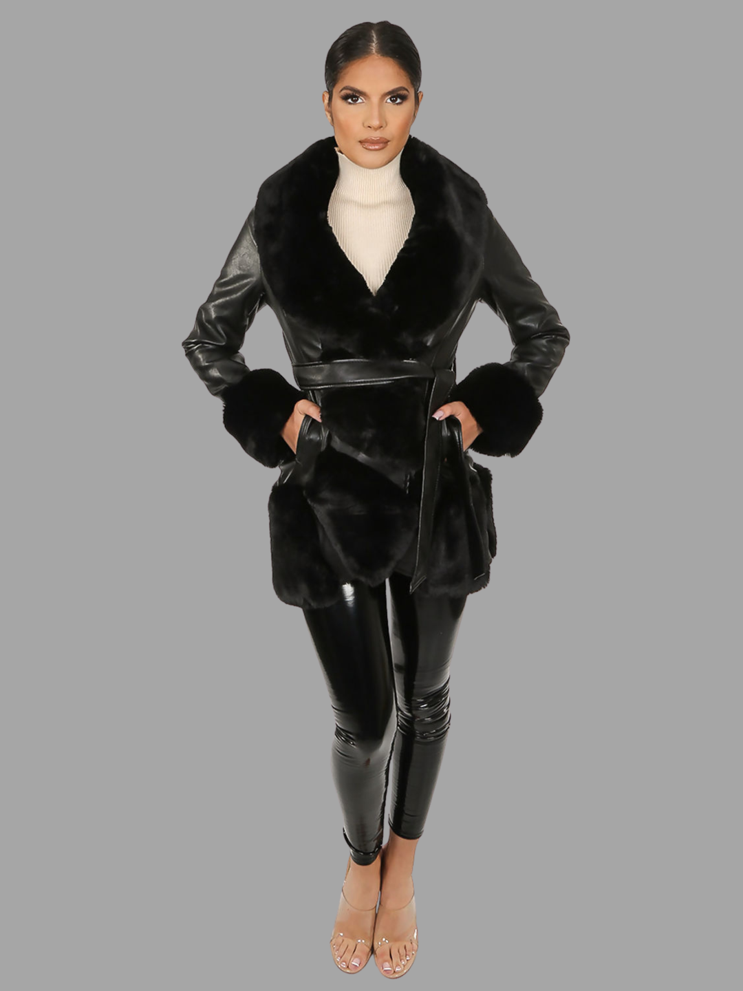 Full length shot. Female model stands in front of a plain grey background. The model wears a black faux leather coat with black faux fur lapel, black faux fur trim and black faux cuffs. The belt has a a faux leather belted waist. The model has tanned skin and black hair that is slicked back. The model faces the camera and places both hands in her pockets, the front faux trim can be seen, including faux fur sleeved cuffs and wet look leggings. 