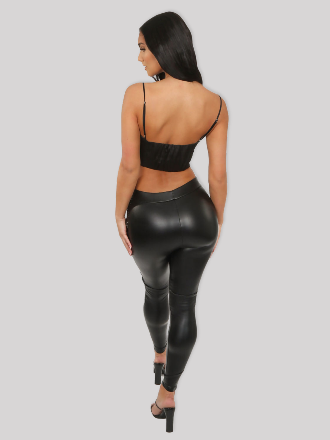 Full length shot. Female model stands in front of a plain grey background, she wears a black faux leather wet look leggings, with a skull embossed side panel. The model has tanned skin and long black hair, she stands with her back to the camera, she wears a black crop top and black mules heels. 