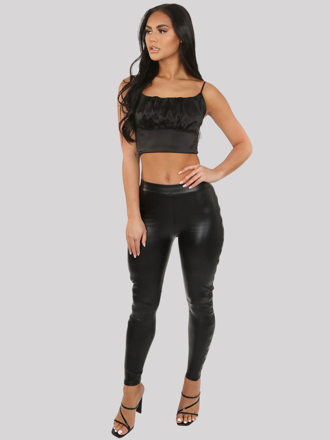 Full length shot. Female model stands in front of a plain grey background, she wears a black faux leather wet look leggings, with a skull embossed side panel. The model has tanned skin and long black hair, she stands slightly to the side and places one hand on her thigh and the other on his hip, she looks off into the camera. She wears a black crop top and black strappy heels.