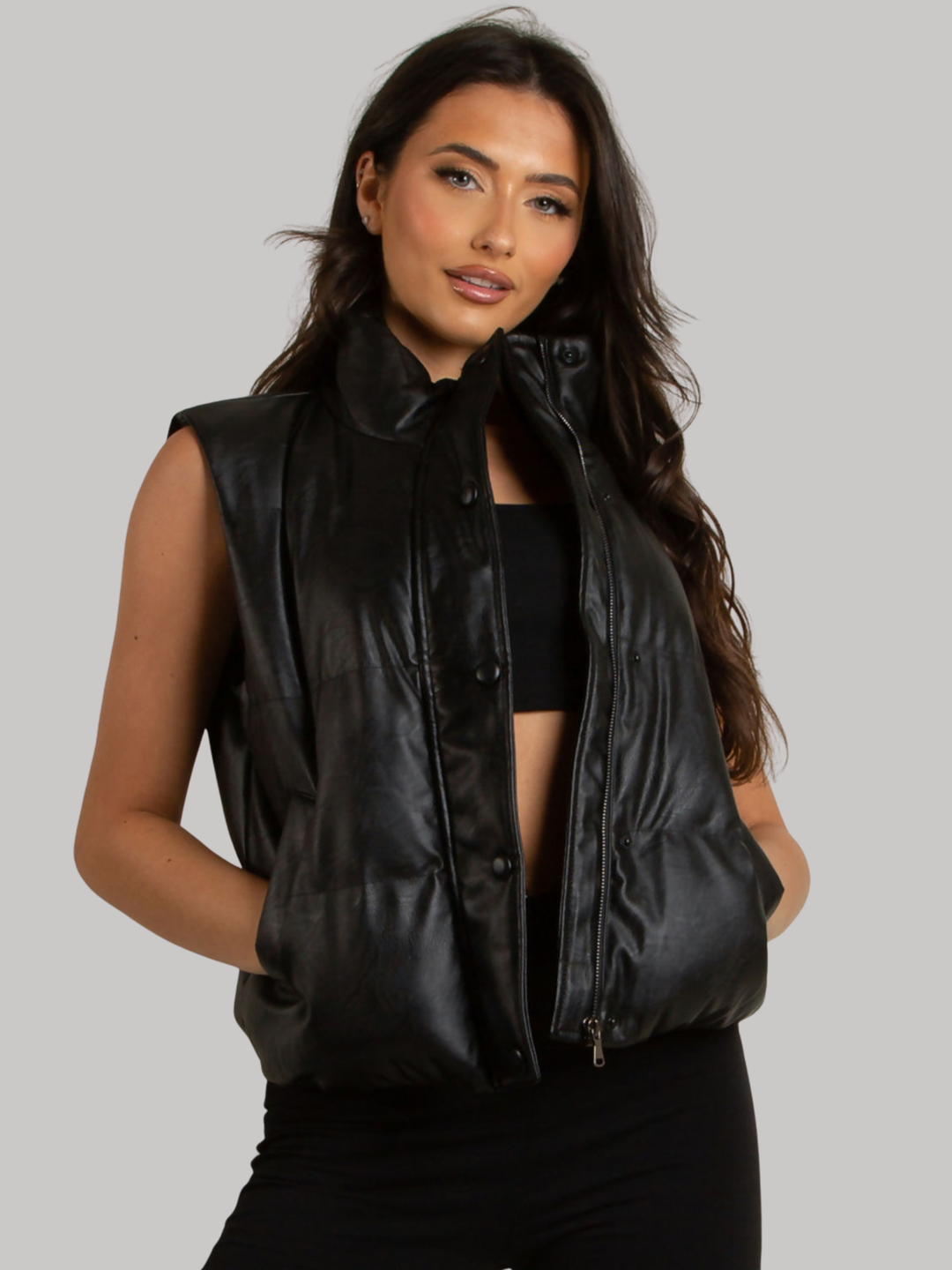Female model stands in front of a plain background. The model wears a black faux leather gillet, with zip and button front fastening with functional side pockets.  The model has tanned skin and long brown hair, she looks directly into the camera, placing her hands in her pockets. 