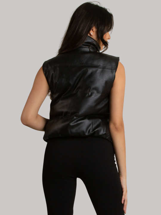 Female model stands in front of a plain background. The model wears a black faux leather gillet, with zip and button front fastening with functional side pockets.  The model has tanned skin and long brown hair,  the model stands with her back to the camera, the back of the puffer can be seen. 
