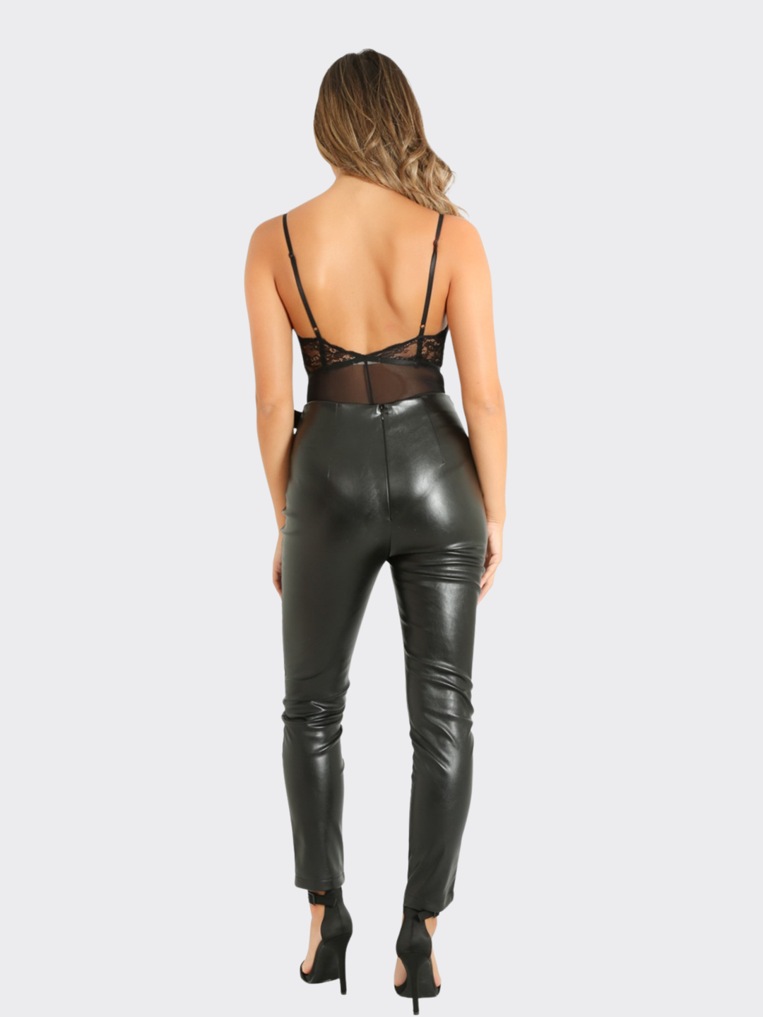Full length shot. Female model stands in front of a blank background, she wears a faux leather trousers with wrap front design, with d buckle fastening. The model stands with her back to the camera, the back of the trousers can be seen.