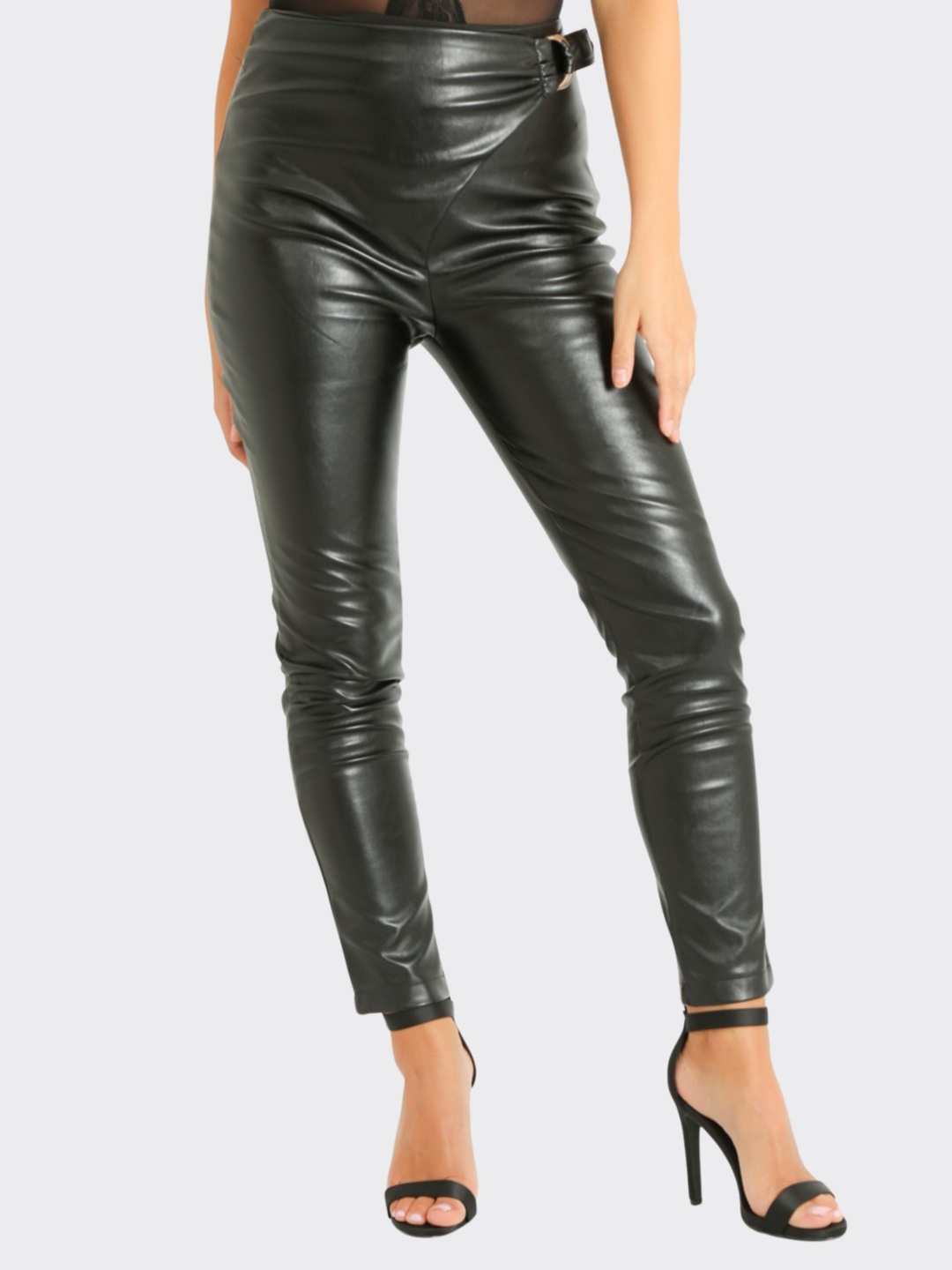 Female model stands in front of a blank background, she wears a faux leather trousers with wrap front design, with d buckle fastening. The model wears black stiletto shoe, she places one hand on her hip and the other behind her back. 