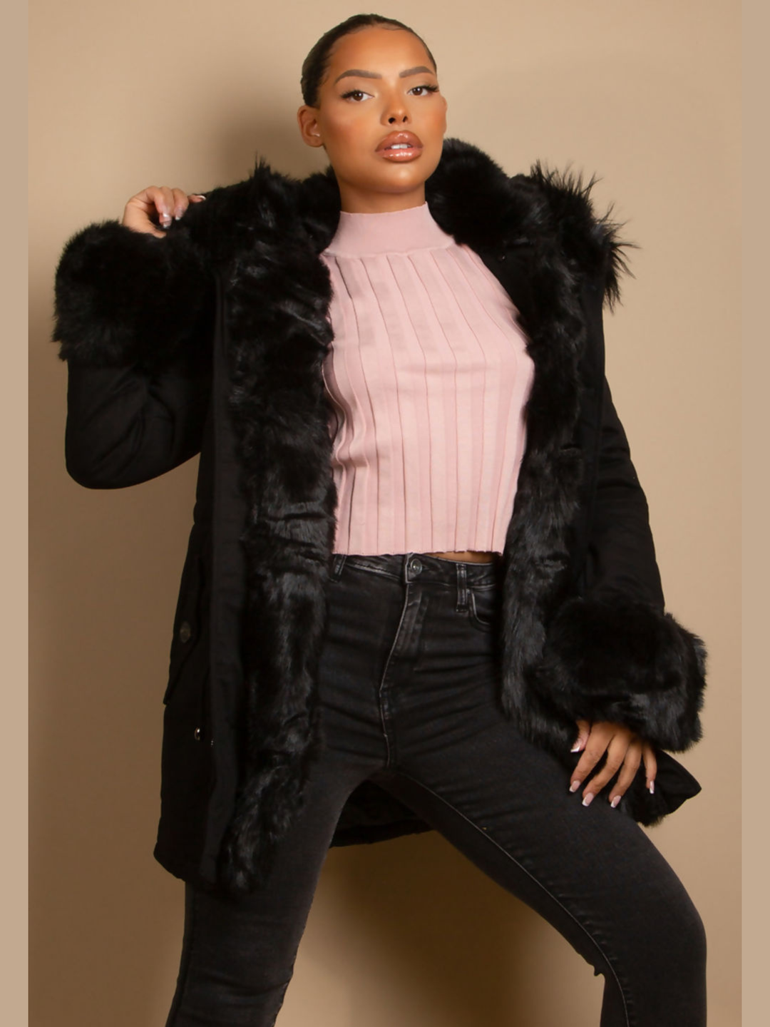 Female model stands in front of a plain background, she wears a black parka coat with front button fastening, faux fur sleeve cuffs, faux fur hood and internal faux fur lining. The model has light brown skin and black hair which is pulled back into a pony tail, she face the camera with the coat open, the inside of the faux fur lining can be seen, she places one hand on her thigh and the other touching the faux fur hood. 