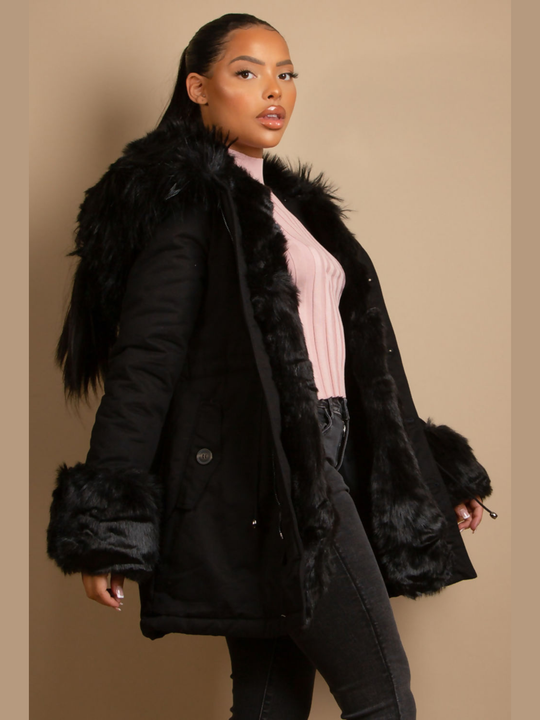 Female model stands in front of a plain background, she wears a black parka coat with front button fastening, faux fur sleeve cuffs, faux fur hood and internal faux fur lining. The model has light brown skin and black hair which is pulled back into a pony tail. The model stands to the side and turns her head to look into the camera. The coat is open, the inside of the faux fur lining can be seen.