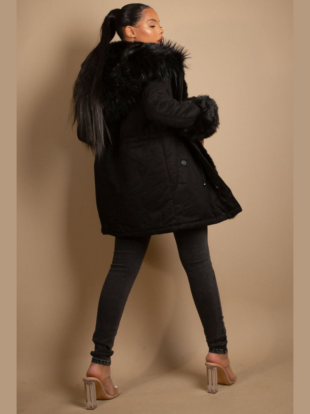 Full length shot. Female model stands in front of a plain background, she wears a black parka coat with front button fastening, faux fur sleeve cuffs, faux fur hood and internal faux fur lining. The model has light brown skin and black hair which is pulled back into a pony tail, she stands with her back to the camera, the back of the parka can be seen, including the faux fur hood and faux fur sleeved cuffs, she wears black skinny fit jeans and perspex heeled shoes.