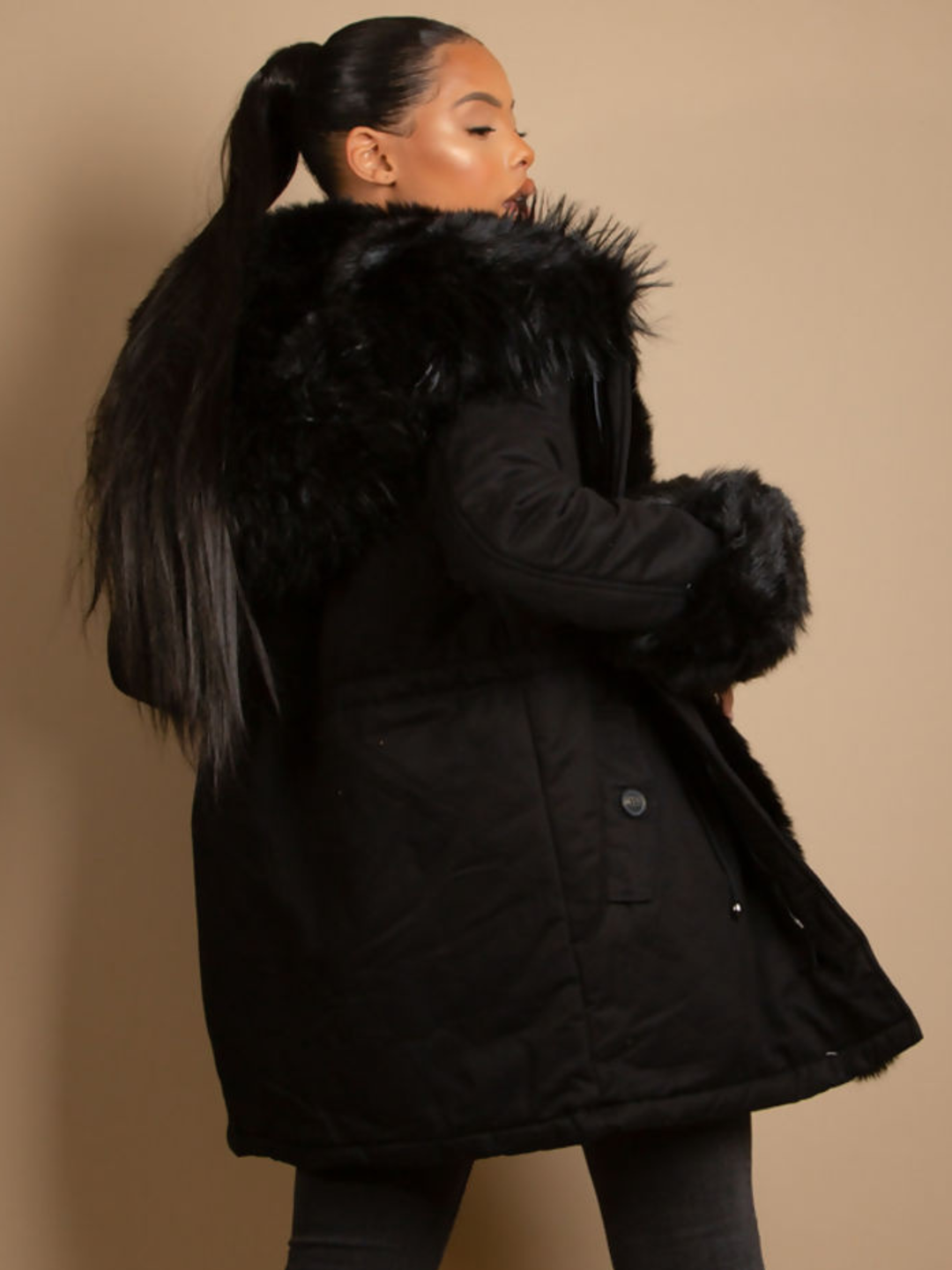 Female model stands in front of a plain background, she wears a black parka coat with front button fastening, faux fur sleeve cuffs, faux fur hood and internal faux fur lining. The model has light brown skin and black hair which is pulled back into a pony tail, she stands with her back to the camera, the back of the parka can be seen, including the faux fur hood and faux fur sleeved cuffs. 