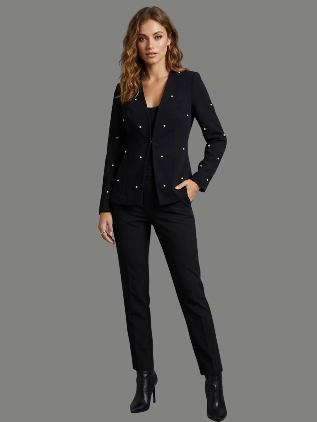 Full length shot. Model stands in front of a plain background, she wears a luxe black lapel-less blazer, with a tailored silhouette, front fastening and  decorative pearls accents  at the front of the blazer and blazer arms. The model has tanned skin, wavy light brown hair and places one hand in her trousers pocket and her hand down her side, she wears black trousers and black heeled ankle boots. 