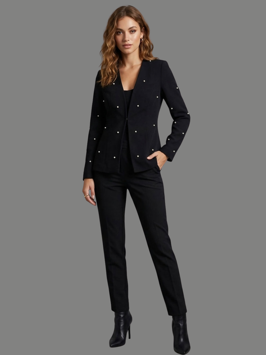 Full length shot. Model stands in front of a plain background, she wears a luxe black lapel-less blazer, with a tailored silhouette, front fastening and  decorative pearls accents  at the front of the blazer and blazer arms. The model has tanned skin, wavy light brown hair and places one hand in her trousers pocket and her hand down her side, she wears black trousers and black heeled ankle boots. 