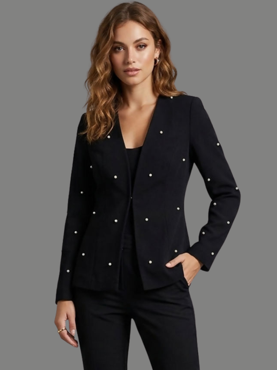 Model stands in front of a plain background, she wears a luxe black lapel-less blazer, with a tailored silhouette, front fastening and  decorative pearls accents  at the front of the blazer and blazer arms. The model has tanned skin, wavy light brown hair and places one hand in her trousers pocket and her hand down her side. 