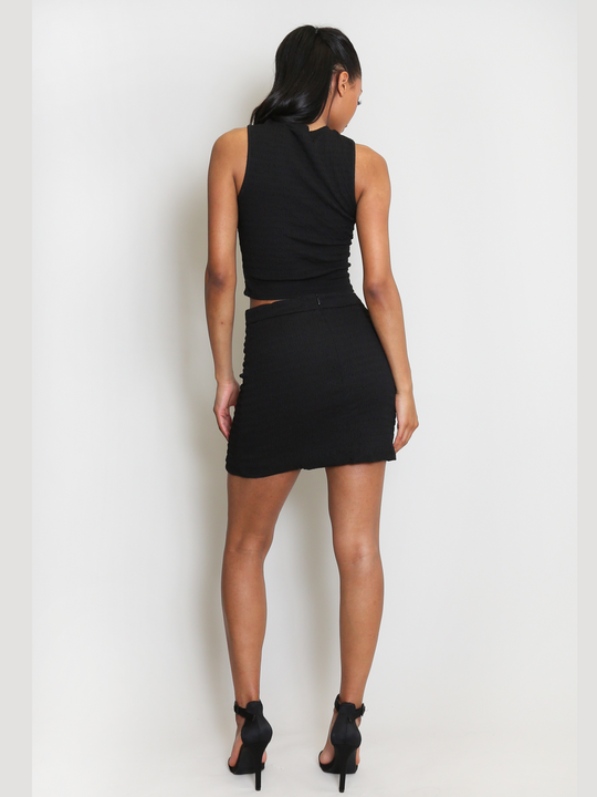 Full length shot. Model wears  luxe black plisse mini skirt with gold military button detailing to the front of the dress. The model has brown skin and long black hair, model stands with her back to the camera, showing the back of the plisse mini skirt.