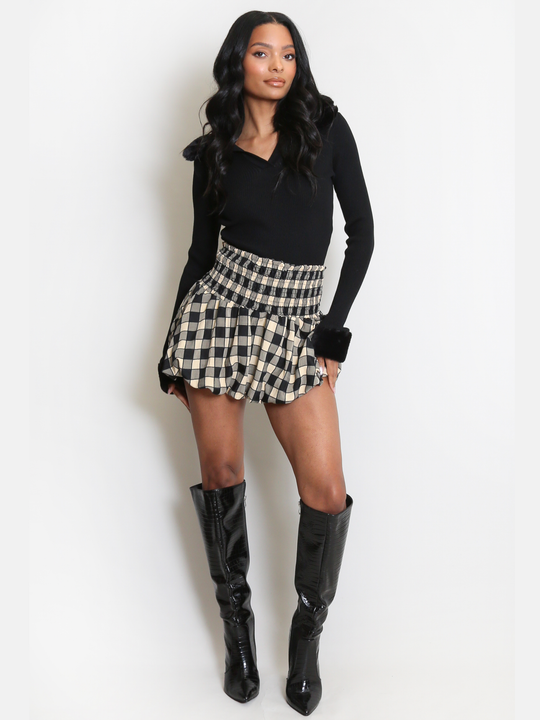 Full length shot. Female model stands in front of a plain background. The model wears a black ribbed jumper with faux fur sleeved cuffs and faux fur collar. The faux fur jumper has a  v neck front. The female model has light brown skin and long black hair. The model faces forward and wears a checkered mini skirt and black knee high boots. 