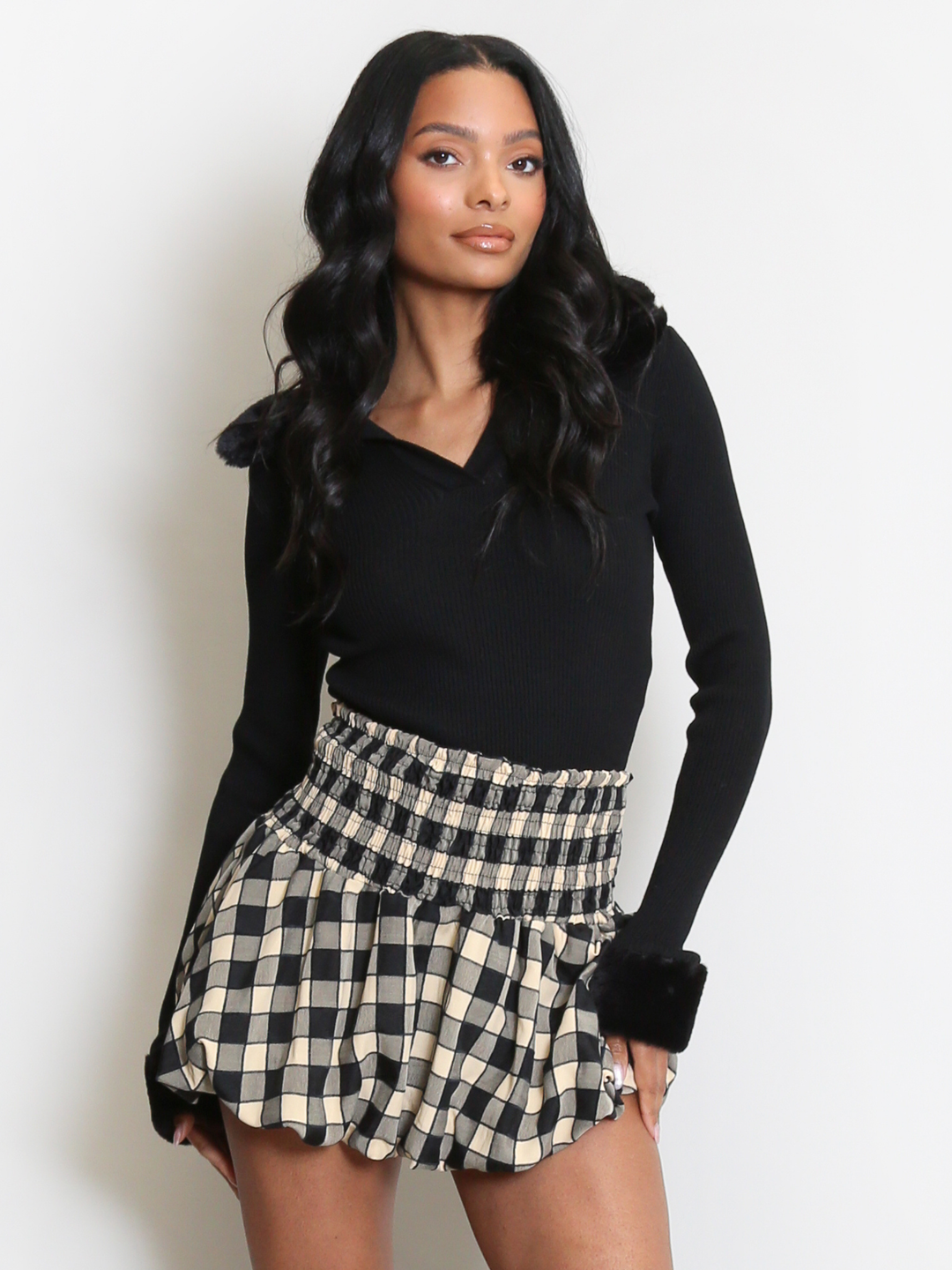 Female model stands in front of a plain background. The model wears a black ribbed jumper with faux fur sleeved cuffs and faux fur collar. The faux fur jumper has a  v neck front. The female model has light brown skin and long black hair. The model faces forward, she places both hands by her sides. 
