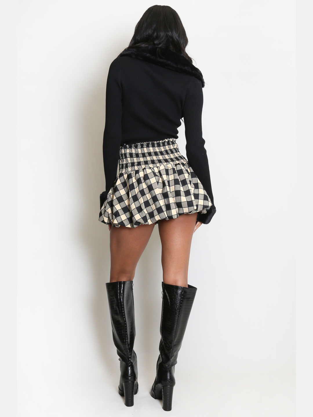 Full length shot. Female model stands in front of a plain background. The model wears a black ribbed jumper with faux fur sleeved cuffs and faux fur collar. The faux fur jumper has a  v neck front. The female model has light brown skin and long black hair, the model stands with her back to the camera, the back of the jumper can be seen, including the faux fur collar and faux fur cuffs. 