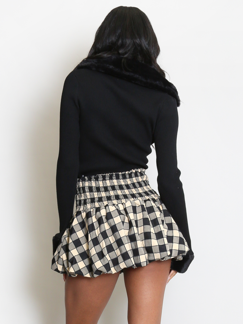 Female model stands in front of a plain background. The model wears a black ribbed jumper with faux fur sleeved cuffs and faux fur collar. The faux fur jumper has a  v neck front. The female model has light brown skin and long black hair, she stands with her back to the camera, the faux fur collar at the back of the jumper can be seen, as well as the faux fur sleeves cuffs. 