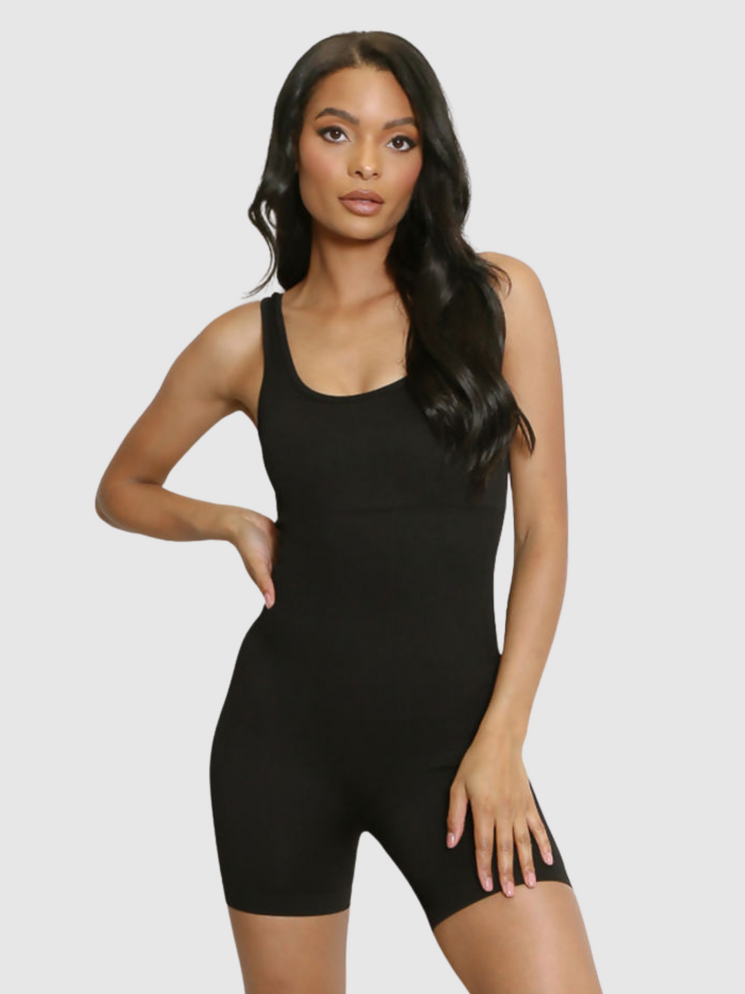 Model wears a black ribbed unitard jumpsuit with a scoop neck, waist control and open back. Model stands with one hand on her hip and one hand on her thigh.