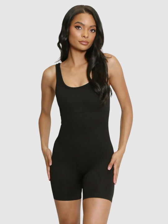 Model wears a black ribbed unitard jumpsuit with a scoop neck, waist control and open back. Model stands with both hands on her hips.