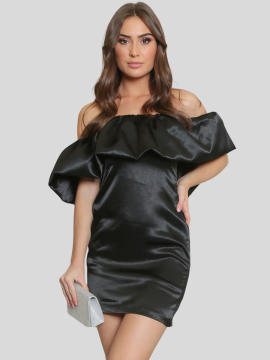 A female model stands before a plain grey background, she wears a black satin mini dress that has an off the shoulder Bardot design. The model faces the camera, she has tanned skin and long brown hair, she places one hand on her hip and holds a clutch in the other. 